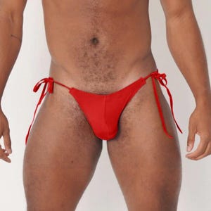 May include: A red thong-style swimsuit with tie closures on the sides. The swimwear is a solid, vibrant red color. The model is standing in front of a white background.