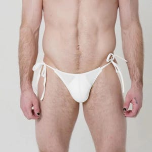 May include: A white, string bikini bottom with tie closures. The swimwear is made of a textured fabric. The model is standing in front of a white background.