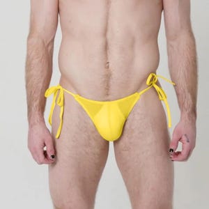 May include: A full-body shot of a person wearing a yellow bikini bottom with tie sides. The swimwear is a bright, solid yellow color. The person is standing against a plain white background.