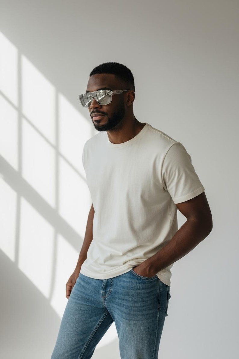 Puede incluir: Una persona con gafas de sol con montura plateada, una camiseta color crema y vaqueros azules. La persona est&aacute; de pie frente a una pared blanca con sombras.