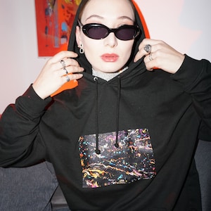 Könnte beinhalten: Ein schwarzer Hoodie mit einem bunten abstrakten Druck auf der Vorderseite. Der Hoodie hat eine Kapuze mit Kordelzug und eine Kängurutasche.