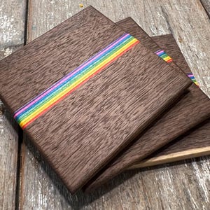May include: Set of four square wooden coasters. Each coaster is a dark brown with a rainbow stripe across the center. The coasters are stacked on a weathered wooden surface.