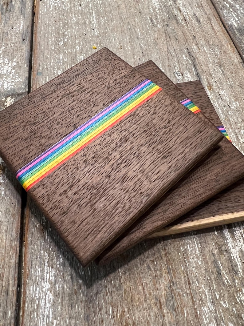 May include: Set of four square wooden coasters. Each coaster is a dark brown with a rainbow stripe across the center. The coasters are stacked on a weathered wooden surface.