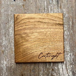 May include: Square wooden coaster with a natural wood grain finish. The coaster has the word "Cartwright" engraved in a cursive font on the lower right corner. The coaster is made of a light brown wood.