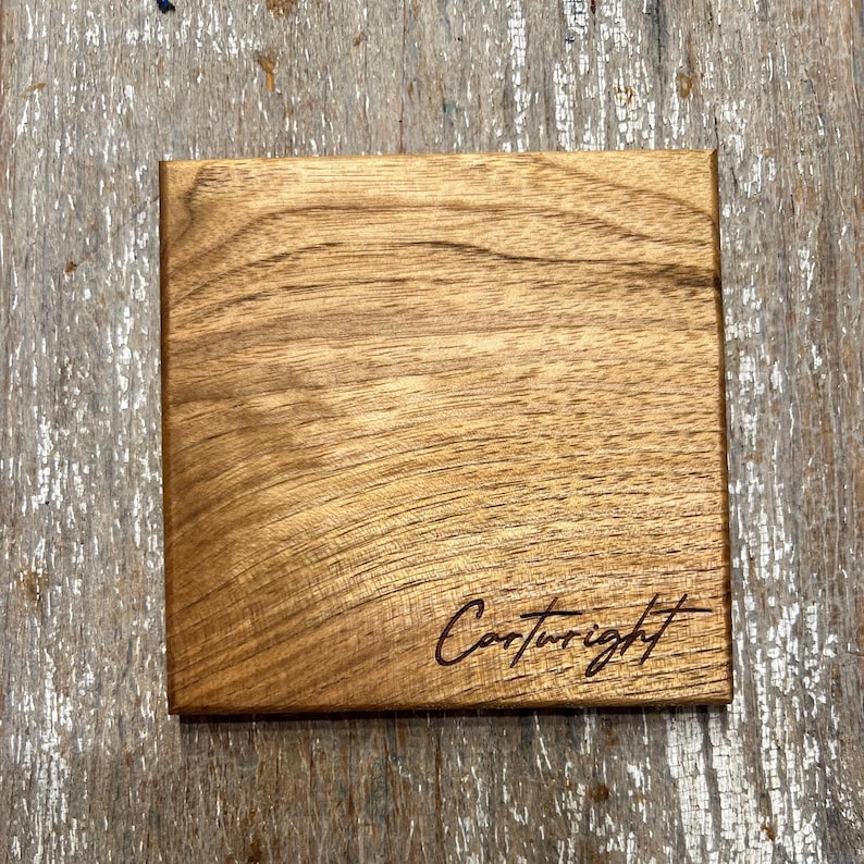 May include: Square wooden coaster with a natural wood grain finish. The coaster has the word "Cartwright" engraved in a cursive font on the lower right corner. The coaster is made of a light brown wood.