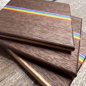 May include: Stack of wooden coasters with a dark brown finish and a rainbow stripe across the center. The coasters have a natural wood grain and a light-colored edge. The rainbow stripe includes red, orange, yellow, green, blue, and purple.