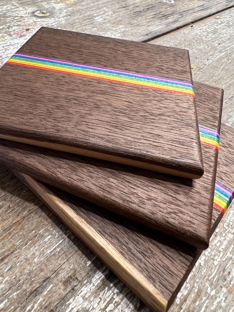 May include: Stack of wooden coasters with a dark brown finish and a rainbow stripe across the center. The coasters have a natural wood grain and a light-colored edge. The rainbow stripe includes red, orange, yellow, green, blue, and purple.