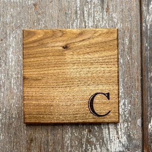May include: Square wooden coaster with a natural wood grain finish. The coaster features a dark, engraved letter "C" in the lower right corner. The wood has a warm, brown tone, and the coaster is set against a rustic wooden background.