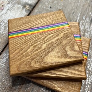 May include: A stack of three square wooden coasters. Each coaster has a natural wood grain finish and a horizontal rainbow stripe across the center. The rainbow stripe includes red, orange, yellow, green, blue, and purple.