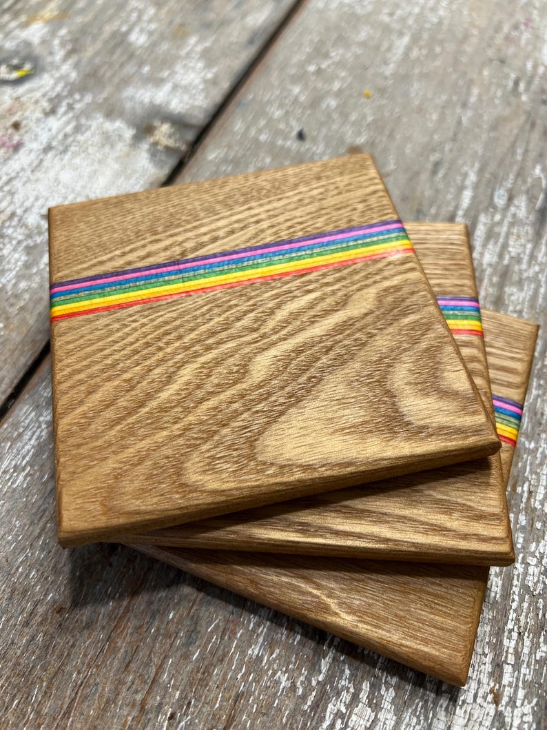 May include: A stack of three square wooden coasters. Each coaster has a natural wood grain finish and a horizontal rainbow stripe across the center. The rainbow stripe includes red, orange, yellow, green, blue, and purple.