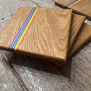 May include: A stack of square wooden coasters with a rainbow stripe design. The coasters are made of light brown wood and have a smooth finish. The rainbow stripe includes red, orange, yellow, green, blue, and purple.