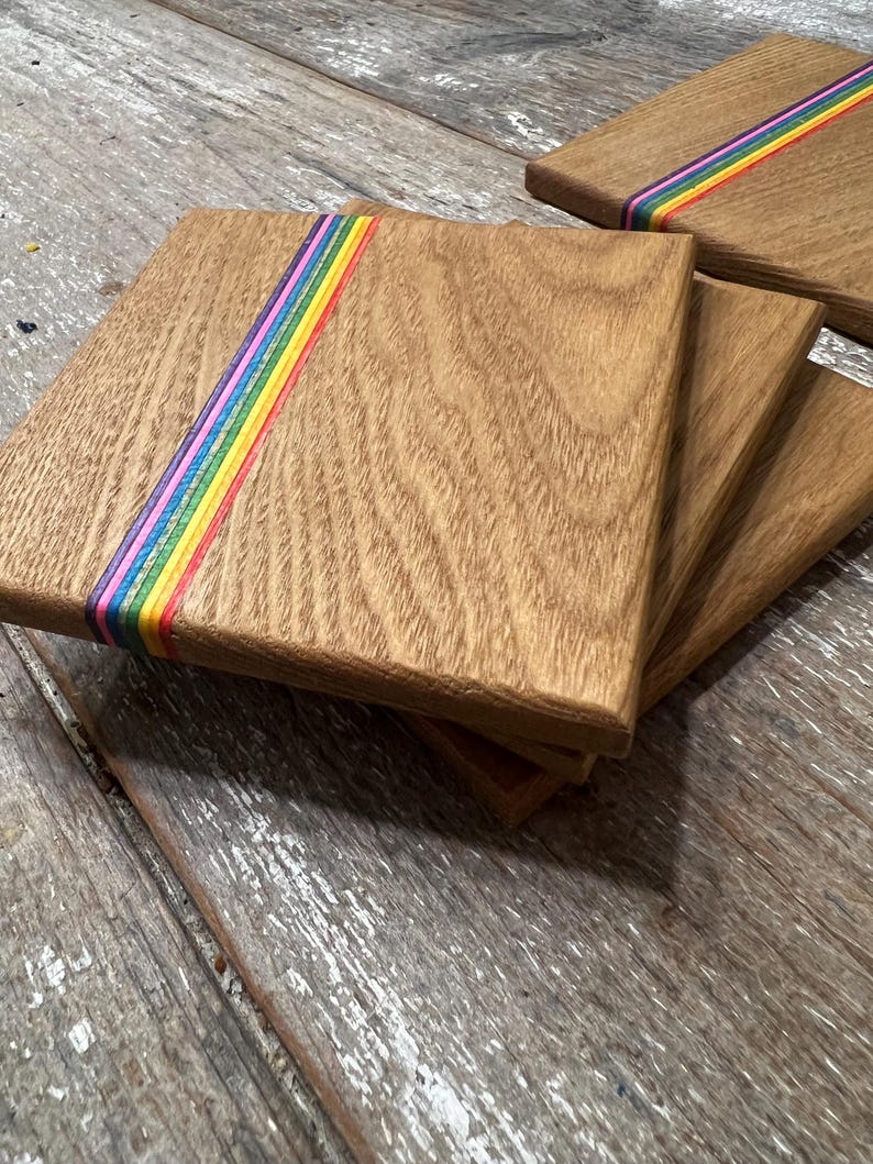 May include: A stack of square wooden coasters with a rainbow stripe design. The coasters are made of light brown wood and have a smooth finish. The rainbow stripe includes red, orange, yellow, green, blue, and purple.