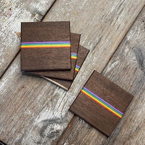 May include: Set of square wooden coasters with a dark brown finish. Each coaster features a horizontal stripe of rainbow colors. The coasters are arranged on a weathered wooden surface.