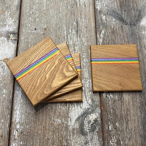 May include: Wooden square coasters with a rainbow stripe design. The coasters are made of light brown wood and feature a horizontal stripe of red, orange, yellow, green, blue, and purple. Several coasters are stacked, and one is separate.