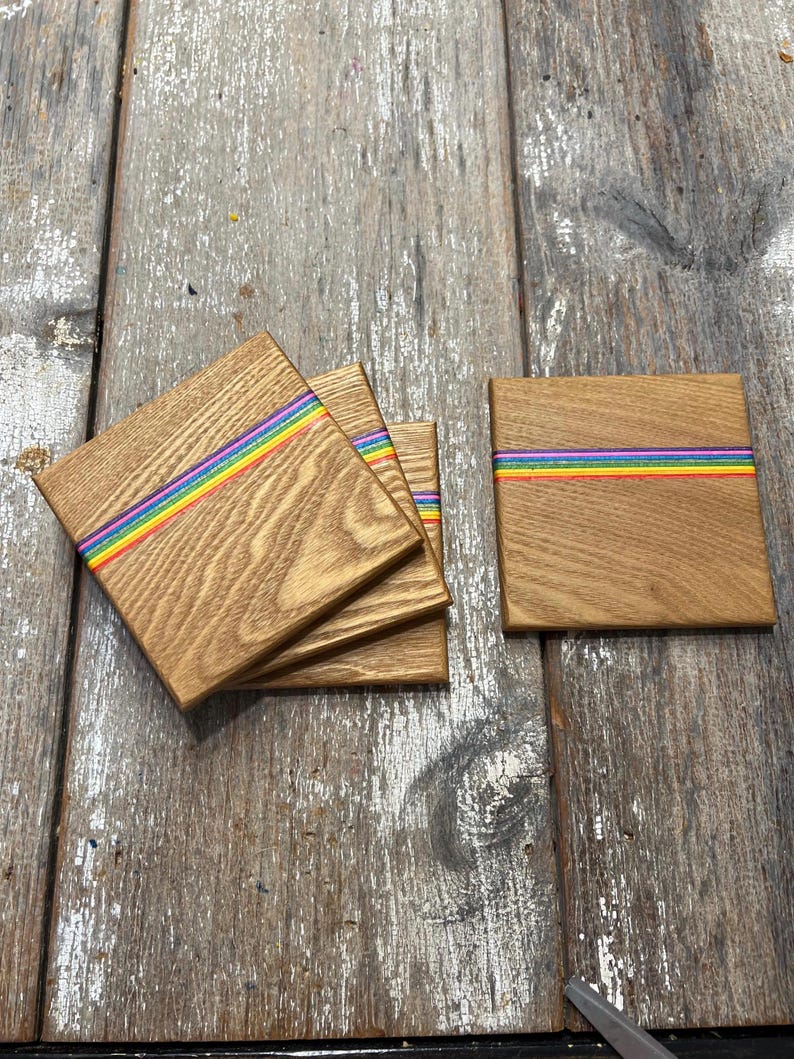 May include: Wooden square coasters with a rainbow stripe design. The coasters are made of light brown wood and feature a horizontal stripe of red, orange, yellow, green, blue, and purple. Several coasters are stacked, and one is separate.