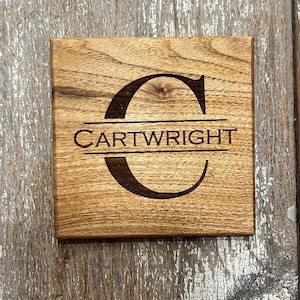 May include: Square wooden coaster with a natural wood grain finish. The coaster features the name "CARTWRIGHT" beneath a large, stylized letter "C". The text and letter are dark brown, contrasting against the lighter wood.