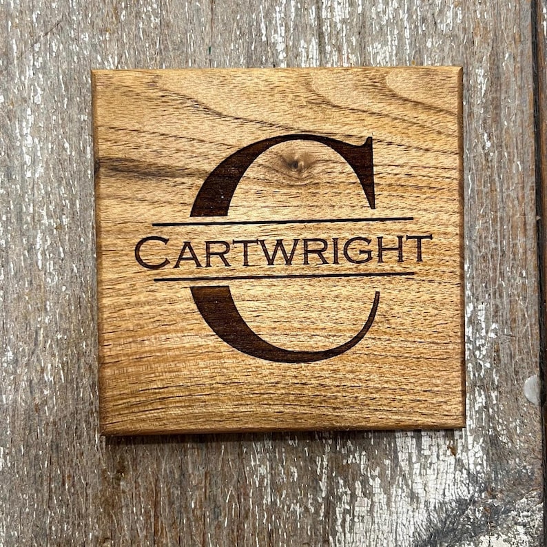 May include: Square wooden coaster with a natural wood grain finish. The coaster features the name "CARTWRIGHT" beneath a large, stylized letter "C". The text and letter are dark brown, contrasting against the lighter wood.