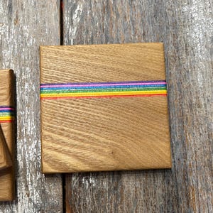 May include: Square wooden coaster with a rainbow stripe design. The coaster is made of light brown wood and features a horizontal band of colorful stripes in red, orange, yellow, green, blue, and purple. The coaster is square and has a smooth surface.