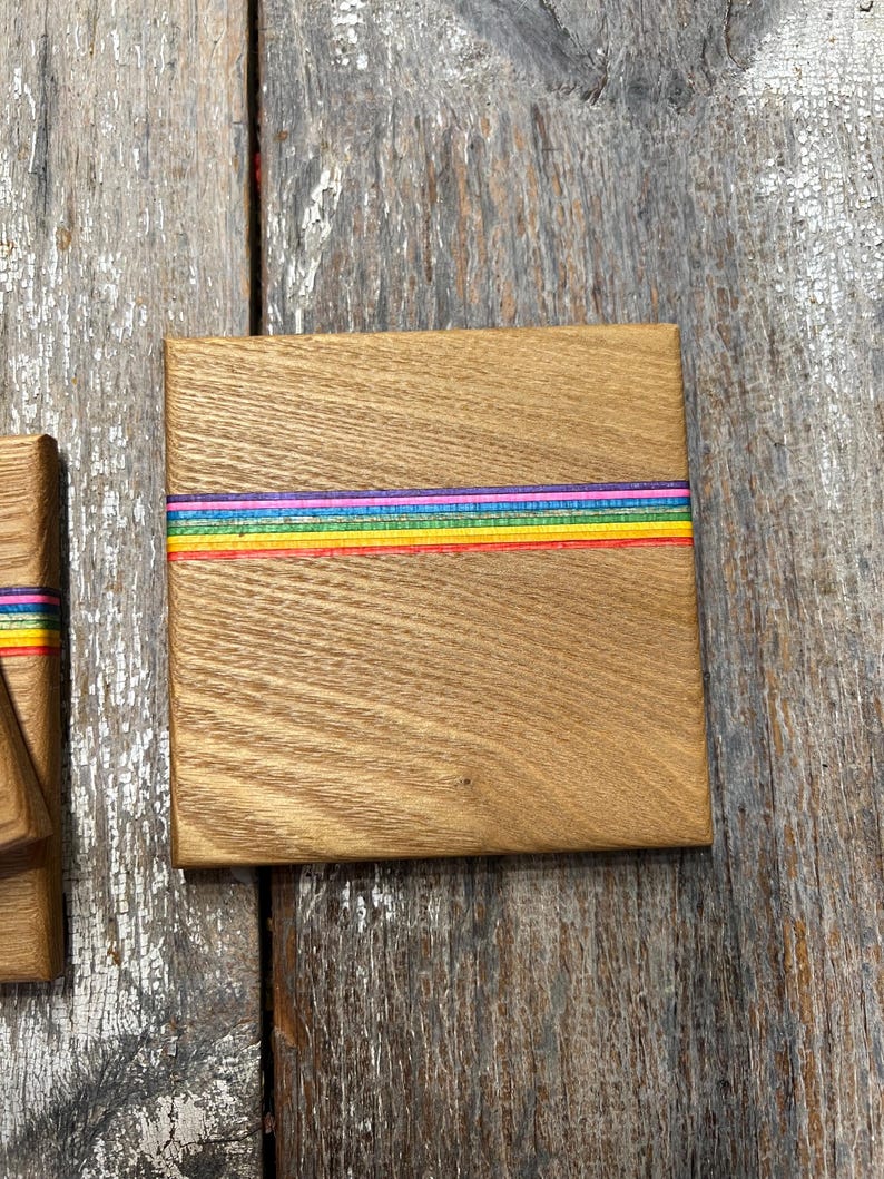 May include: Square wooden coaster with a rainbow stripe design. The coaster is made of light brown wood and features a horizontal band of colorful stripes in red, orange, yellow, green, blue, and purple. The coaster is square and has a smooth surface.