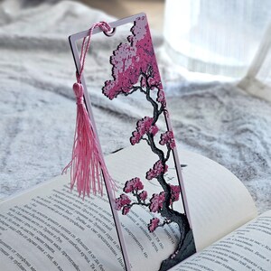 May include: A decorative bookmark featuring a pink cherry blossom tree design. The bookmark has a pink tassel and is placed in an open book. The tree has a black trunk and branches with pink flowers.
