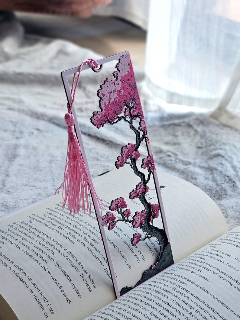 May include: A decorative bookmark featuring a pink cherry blossom tree design. The bookmark has a pink tassel and is placed in an open book. The tree has a black trunk and branches with pink flowers.