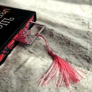 May include: A pink metal bookmark with a decorative floral design is inserted into a book. The bookmark has a matching pink tassel. The book has a black cover with red floral accents.