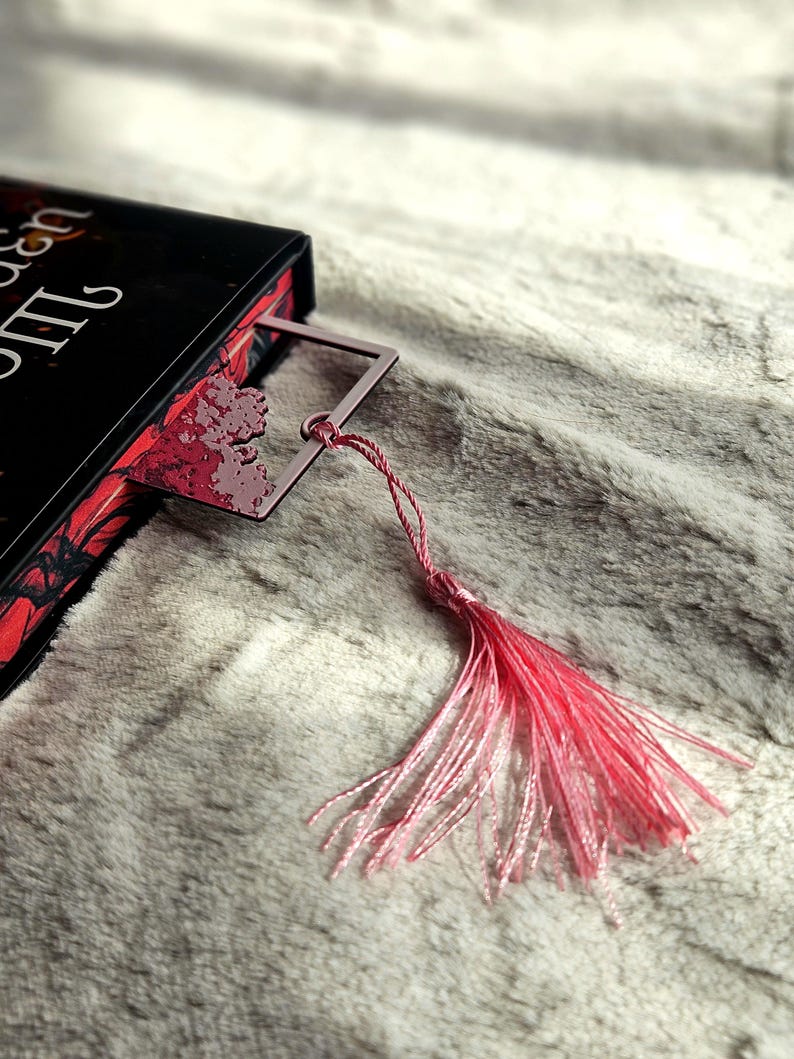 May include: A pink metal bookmark with a decorative floral design is inserted into a book. The bookmark has a matching pink tassel. The book has a black cover with red floral accents.