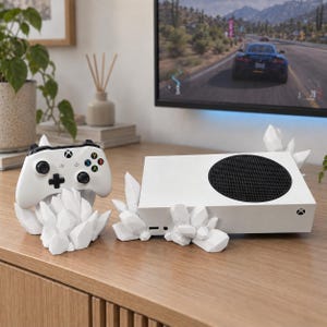 May include: A white Xbox Series S game console and a matching white wireless controller, both displayed with custom white, geometric, crystal-shaped stands. The console features a prominent black circular vent, and the controller has standard buttons.