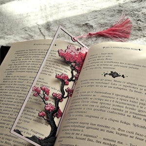 May include: A decorative bookmark featuring a laser-cut cherry blossom tree design in pink and black, placed within the pages of an open book. A pink tassel hangs from the top. The book is open to a page with text.
