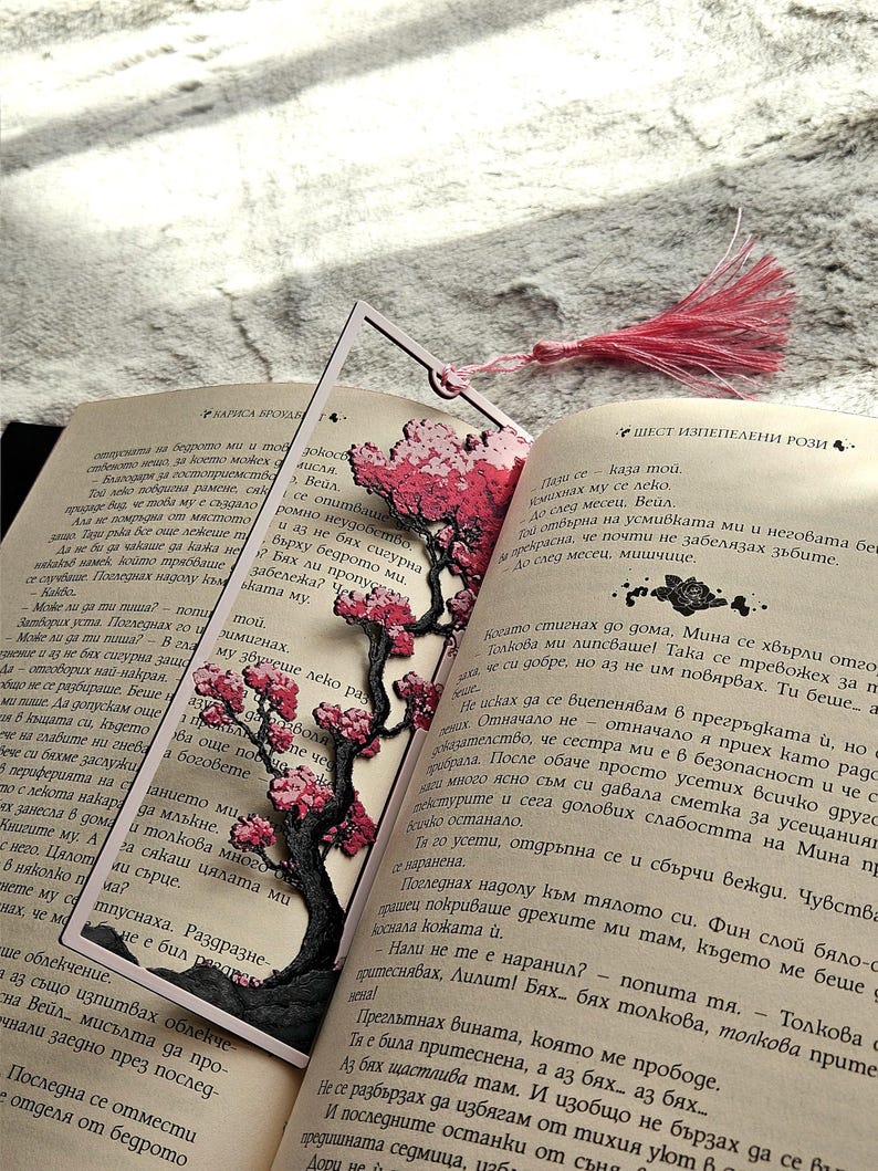 May include: A decorative bookmark featuring a laser-cut cherry blossom tree design in pink and black, placed within the pages of an open book. A pink tassel hangs from the top. The book is open to a page with text.