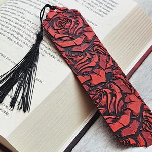 May include: A rectangular bookmark featuring a raised, textured pattern of red roses against a black background. The bookmark is finished with a thin black cord tassel attached to the top, creating a contrast between the vibrant red and deep black.