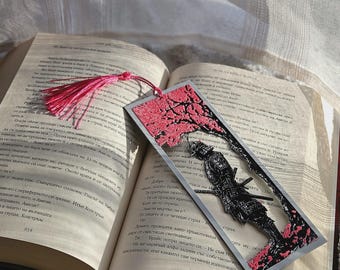 May include: A rectangular bookmark featuring a black silhouette of a samurai standing under a pink cherry blossom tree. The bookmark has a silver-toned border and a pink tassel attached to the top. It is resting on an open book with printed text.