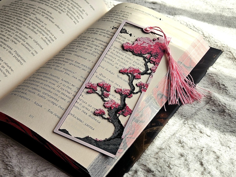 May include: A decorative bookmark featuring a detailed tree design with pink blossoms and a pink tassel. The bookmark is placed in an open book, with text visible on the pages. The bookmark has a black border.