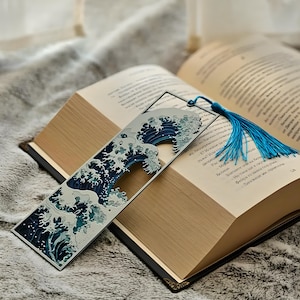 May include: A metal bookmark featuring a stylized wave design in shades of blue and white, with a blue tassel. The bookmark is placed in an open book with gold-toned pages, resting on a soft, gray surface.