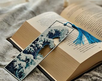 May include: A metal bookmark featuring a stylized wave design in shades of blue and white, with a blue tassel. The bookmark is placed in an open book with gold-toned pages, resting on a soft, gray surface.