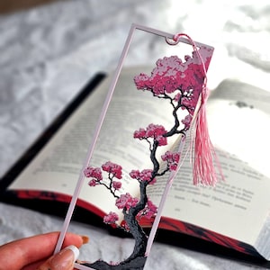 Puede incluir: Marcapáginas transparente con diseño de cerezo en flor. El árbol tiene un tronco gris oscuro y flores rosas. El marcapáginas tiene un borde rosa y una borla rosa. Se encuentra sobre un libro abierto.