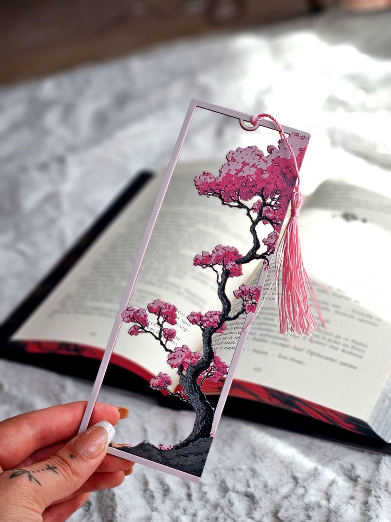 May include: A pink and clear bookmark featuring a cherry blossom tree design. The tree has a dark gray trunk and branches with pink blossoms. A pink tassel hangs from the top of the bookmark, which is held over an open book.
