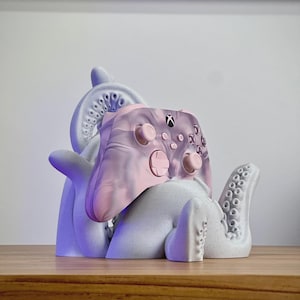 May include: A light grey octopus-shaped controller stand with textured suction cups holding a pink and grey marbled wireless gaming controller. The controller features a central Xbox logo, two analogue sticks, a directional pad, and four action buttons.