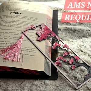May include: A decorative bookmark featuring a pink cherry blossom tree design with a pink tassel. The bookmark is rectangular with a framed design and is placed in an open book. The text "AMS NOT REQUIRED" is visible.