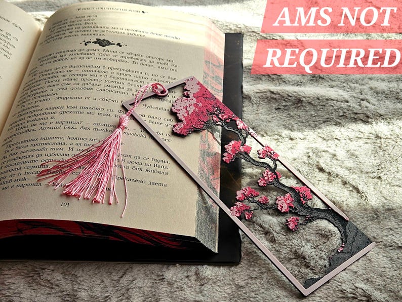 May include: A decorative bookmark featuring a pink cherry blossom tree design with a pink tassel. The bookmark is rectangular with a framed design and is placed in an open book. The text "AMS NOT REQUIRED" is visible.