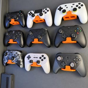 May include: A collection of nine various video game controllers mounted on a dark gray wall using bright orange plastic wall mounts. The controllers include black, white, and camouflage designs, each held securely by the orange support brackets.