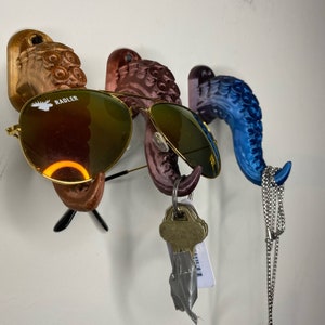 Puede incluir: Un soporte de pared para gafas de sol y llaves con tres ganchos decorativos impresos en 3D en tonos marrón, morado y azul. Gafas de sol con montura dorada y llaves cuelgan de los ganchos. Un collar plateado cuelga del gancho azul.