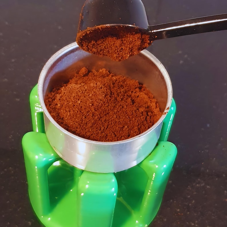 K&ouml;nnte beinhalten: Ein Kaffeekorb aus silberfarbenem Metall, gef&uuml;llt mit dunkelbraunem Kaffeepulver, der in einem hellgr&uuml;nen Kunststoffst&auml;nder steht. Ein schwarzer Kunststoffl&ouml;ffel befindet sich &uuml;ber dem Korb und enth&auml;lt eine kleine Portion des Kaffeepulvers.