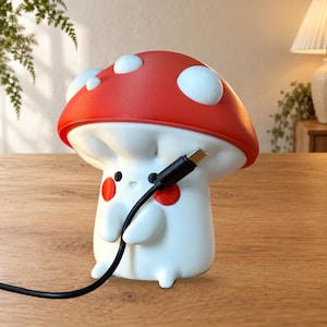 May include: A white mushroom-shaped lamp with a red cap featuring white spots. The lamp has a cartoon face with small black eyes and red cheeks. It is held in a standing position with a black USB charging cable plugged into the front side.