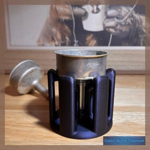 May include: A metal coffee maker component with a dark blue, almost black, 3D-printed holder. The text "Color- Burnt Titanium" is visible at the bottom. The coffee maker is on a wooden surface.