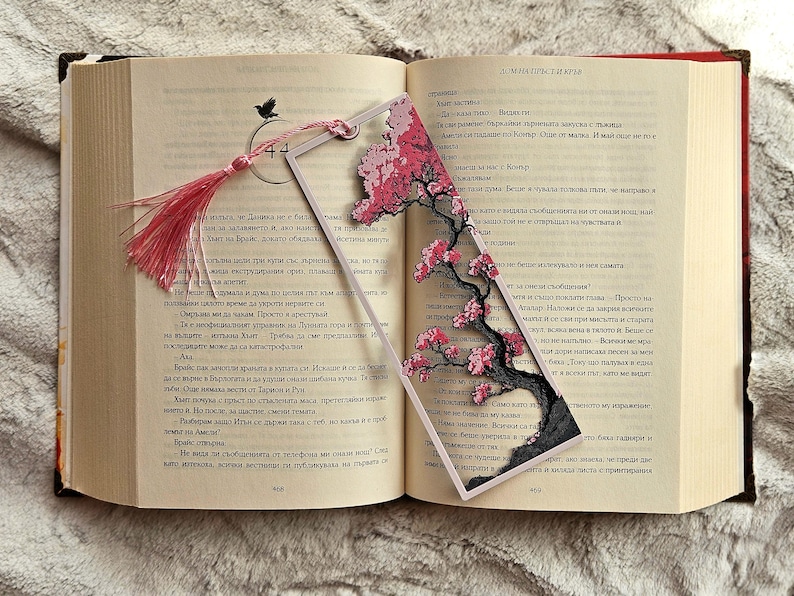 May include: A decorative bookmark featuring a cherry blossom design with pink flowers and a black tree trunk. The bookmark has a pink tassel and is placed in an open book.