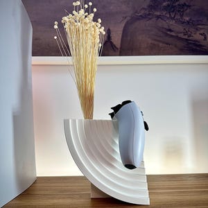 May include: A white, semi-circular decorative table with a ridged design, holding a bundle of dried beige flowers and a light blue and black game controller. The table is on a wooden surface, with a painting in the background.