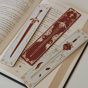 May include: Three rectangular bookmarks resting on an open book. Two are light gray with gold-toned sword illustrations and ornate filigree borders. The center bookmark is deep red with a gold-toned sword design and intricate decorative accents.