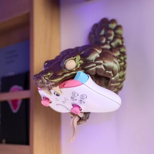 May include: A wall-mounted, bronze-colored snake sculpture with textured scales and fangs, holding a white Xbox wireless controller with pink thumbsticks and a light blue bumper button in its open mouth, featuring a protruding snake tongue.