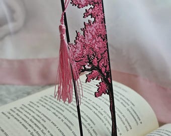 May include: A rectangular black bookmark featuring a laser-cut design of a tree with textured pink blossoms. The bookmark includes a pink tassel attached to the top corner with a thin cord, resting vertically against an open book with printed text.
